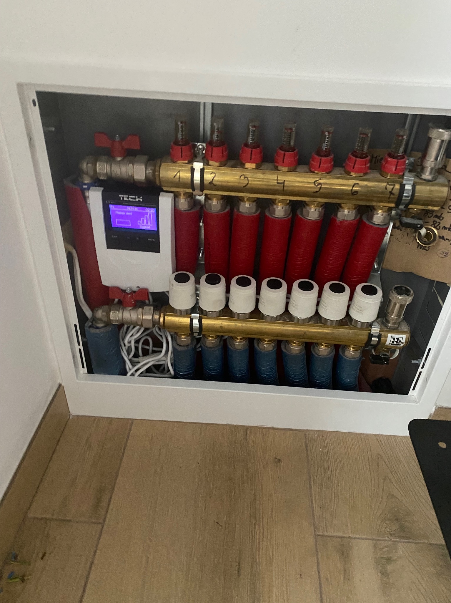 Rozdzielacz ogrzewania podłogowego TECH w szafce, z widocznymi rurami w kolorach czerwonym i niebieskim, oraz elektronicznym sterownikiem. Instalacja grzewcza.