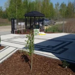 Ułożona kostka brukowa w geometryczny wzór z obrzeżami, rabaty z korą, młode drzewka i altana w tle.