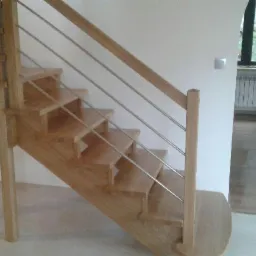 Drewniane schody z metalowymi poręczami w jasnym wnętrzu, widoczne elementy wykończeniowe i okno z radiatorem w tle.