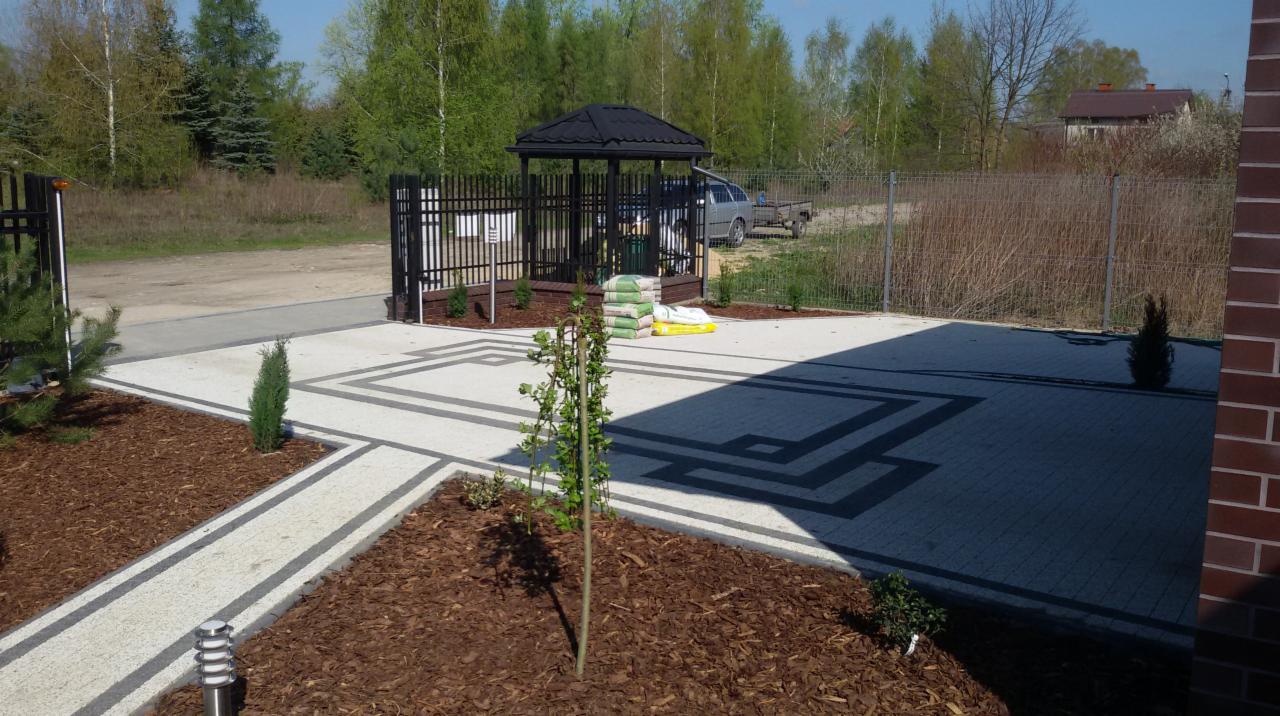 Ułożona kostka brukowa w geometryczny wzór z obrzeżami, rabaty z korą, młode drzewka i altana w tle.