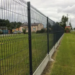 Ciemnoszare ogrodzenie panelowe z podmurówką betonową, widok z boku na tle zieleni i zabudowań. Słoneczny dzień, częściowo zachmurzone niebo.