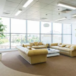 Jasny, przestronny hol biurowy z panoramicznymi oknami, beżowymi sofami i roślinami w donicach. Wykładzina w odcieniach brązu. Nowoczesne oświetlenie sufitowe.