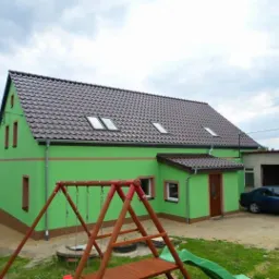 Jaskrawozielony dom jednorodzinny z brązowym dachem i elementami wykończeniowymi, widoczny plac zabaw i garaż z samochodem w tle.