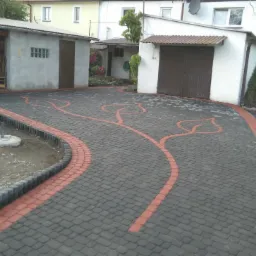 Ułożona kostka brukowa w kolorze szarym z czerwonymi akcentami, tworząca wzór liści. Widoczny fragment ogrodu i budynki w tle. Całość tworzy estetyczny podjazd.
