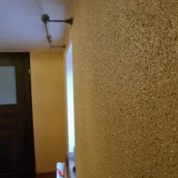 Błyszcząca, złota ściana z wyraźną fakturą w odcieniach beżu i brązu. Widoczne narzędzia malarskie przy ścianie,  oświetlenie i fragment drzwi.