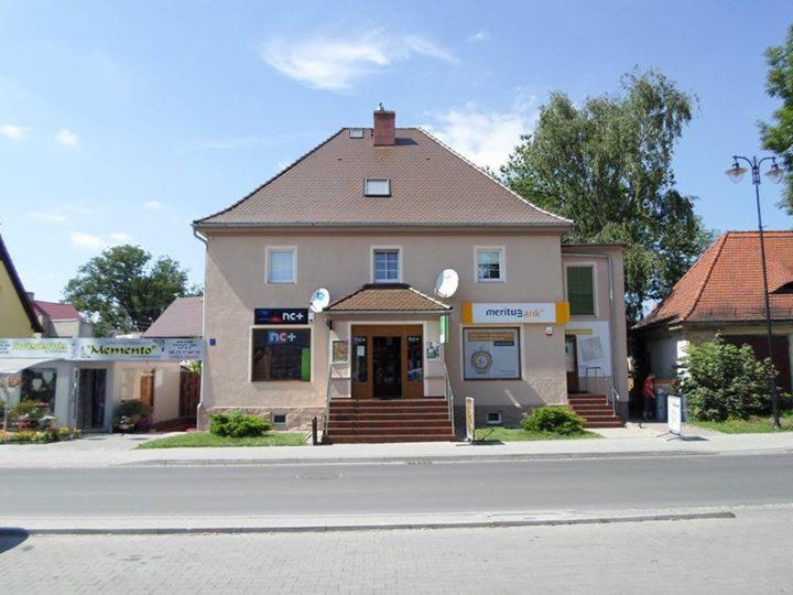 Dwupiętrowy budynek z beżową elewacją, brązowym dachem i schodami wejściowymi. Widoczne szyldy firmowe i anteny satelitarne. Ujęcie z poziomu ulicy w słoneczny dzień.