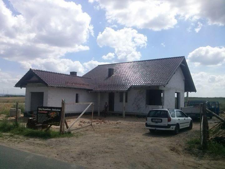 Niedokończony dom murowany z ciemną dachówką w słoneczny dzień. Widoczna tablica informacyjna, samochód osobowy i ciężarowy. W tle pole i błękitne niebo z chmurami.