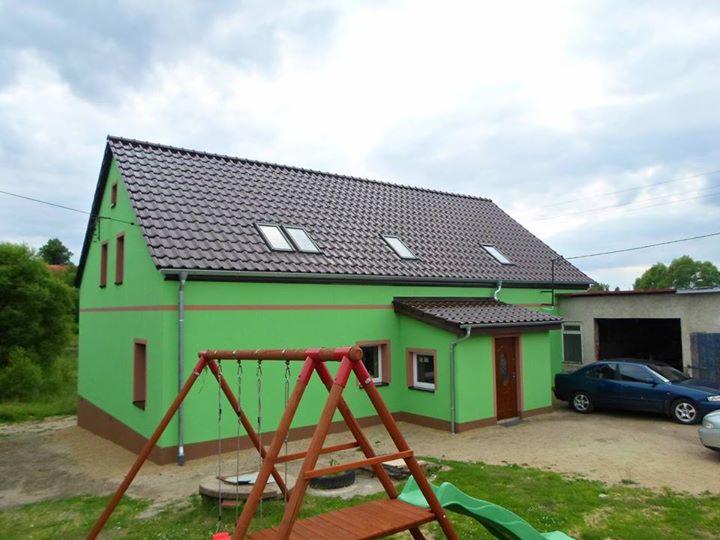 Jaskrawozielony dom jednorodzinny z brązowym dachem i elementami wykończeniowymi, widoczny plac zabaw i garaż z samochodem w tle.