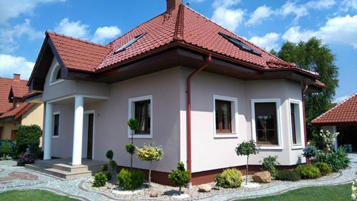 Jasny dom jednorodzinny z czerwoną dachówką, widok z zewnątrz, z zadbanym ogrodem i ścieżką z kostki brukowej. Widoczne okna z brązowymi ramami i białe kolumny przy wejściu.