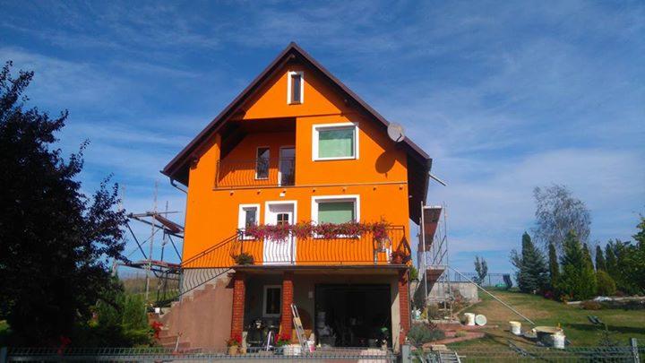 Pomarańczowy dom jednorodzinny z balkonem, po wykonaniu elewacji. Widoczne rusztowania i elementy ogrodu. Słoneczny dzień, błękitne niebo.