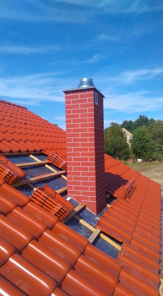 Ceglany komin z metalowym zakończeniem na dachu z czerwonej dachówki, częściowo w trakcie układania. Widoczne drewniane elementy konstrukcji dachu i błękitne niebo.