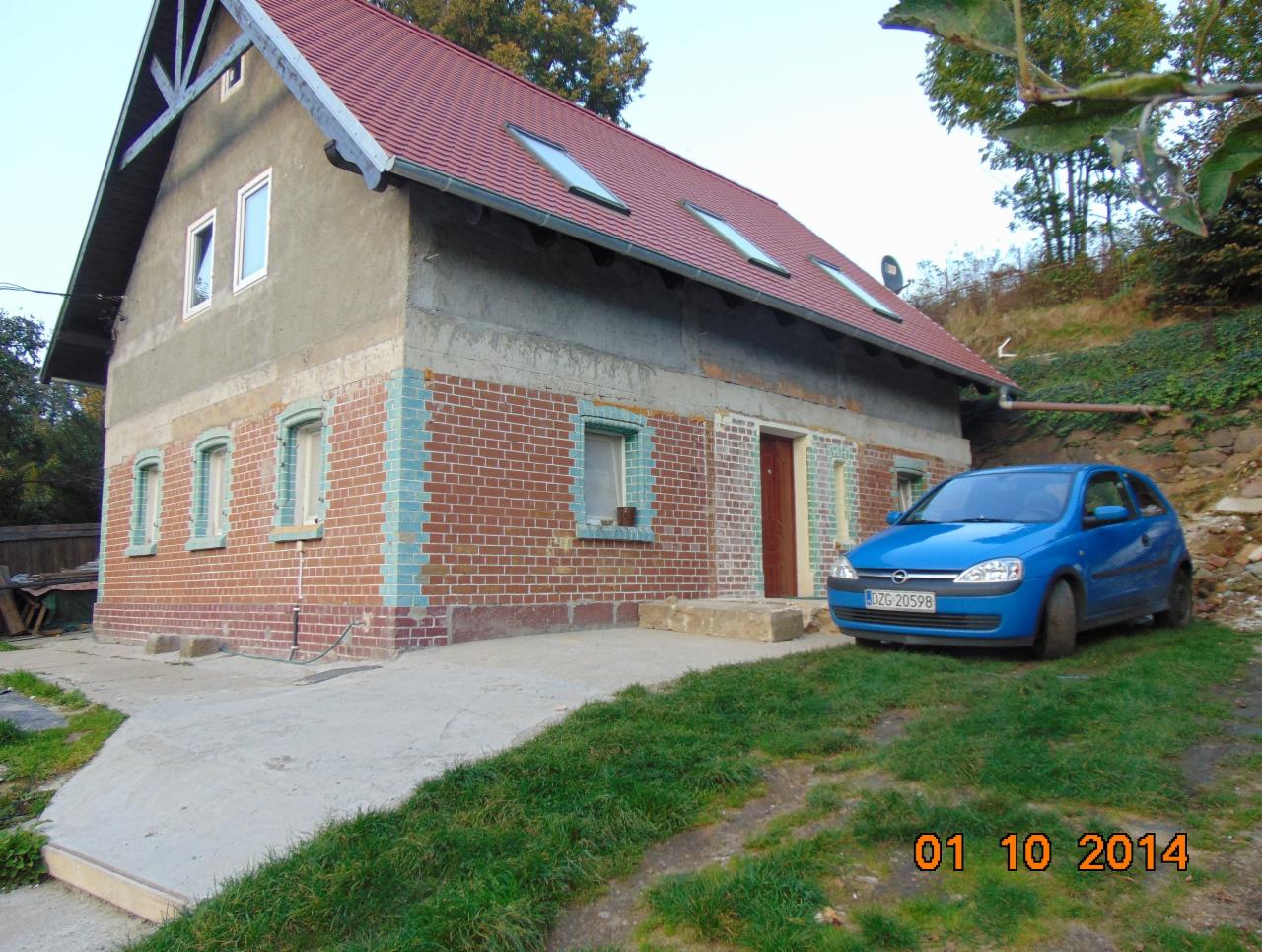 Niedokończony dom z czerwonej cegły i szarym tynkiem, widoczne okna dachowe, zaparkowany niebieski samochód. Budowa w trakcie prac murarskich i tynkarskich.