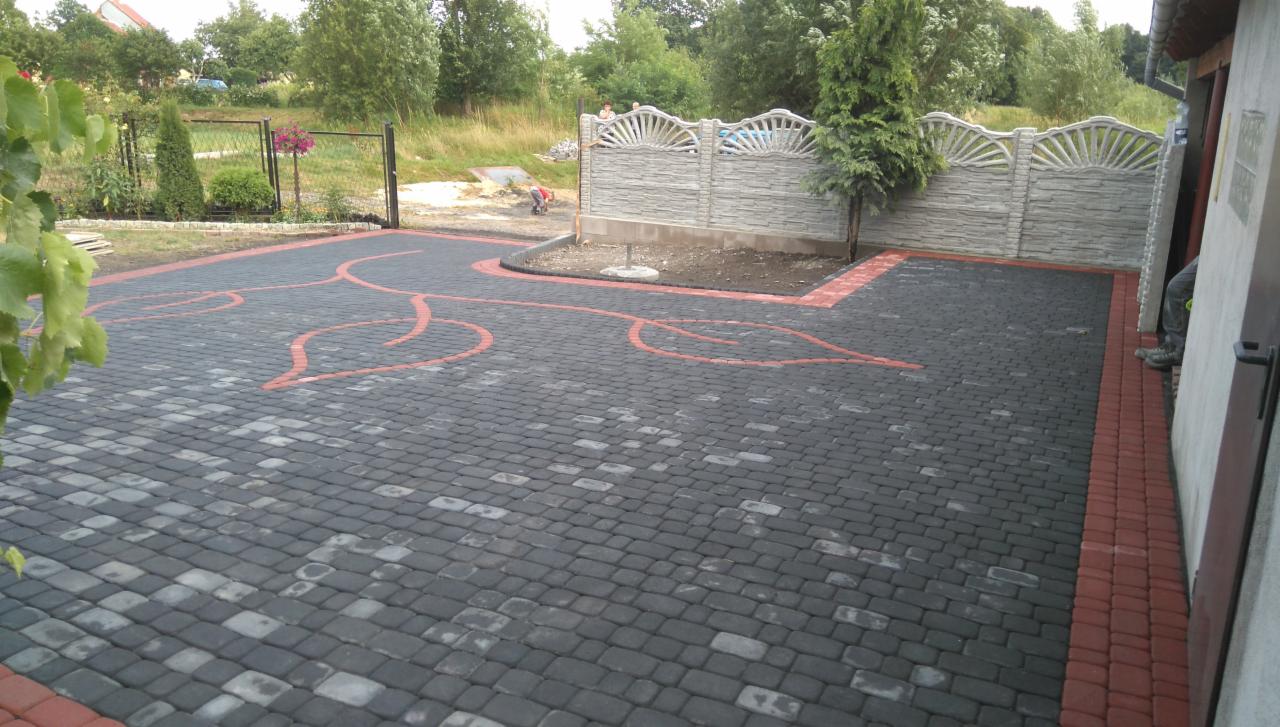 Ułożona kostka brukowa w kolorze grafitowym z czerwonym wzorem liści, obrzeże z czerwonej kostki. W tle ogrodzenie z betonu architektonicznego i zieleń.