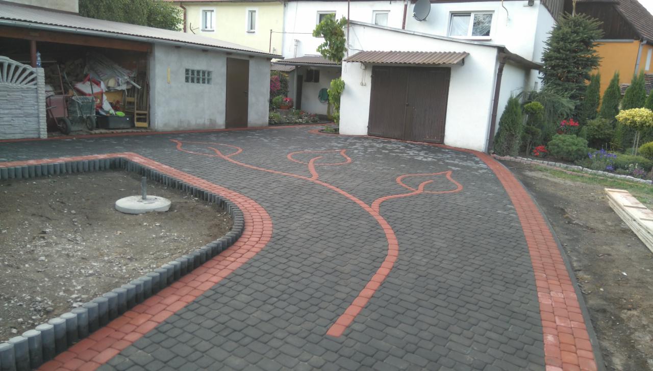 Ułożona kostka brukowa w kolorze szarym z czerwonymi akcentami, tworząca wzór liści. Widoczny fragment ogrodu i budynki w tle. Całość tworzy estetyczny podjazd.