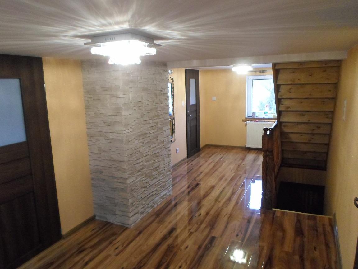 Jasne wnętrze z drewnianą podłogą, ścianą z kamienia dekoracyjnego i schodami z surowego drewna. Widoczne drzwi i okno z grzejnikiem. Oświetlenie sufitowe.