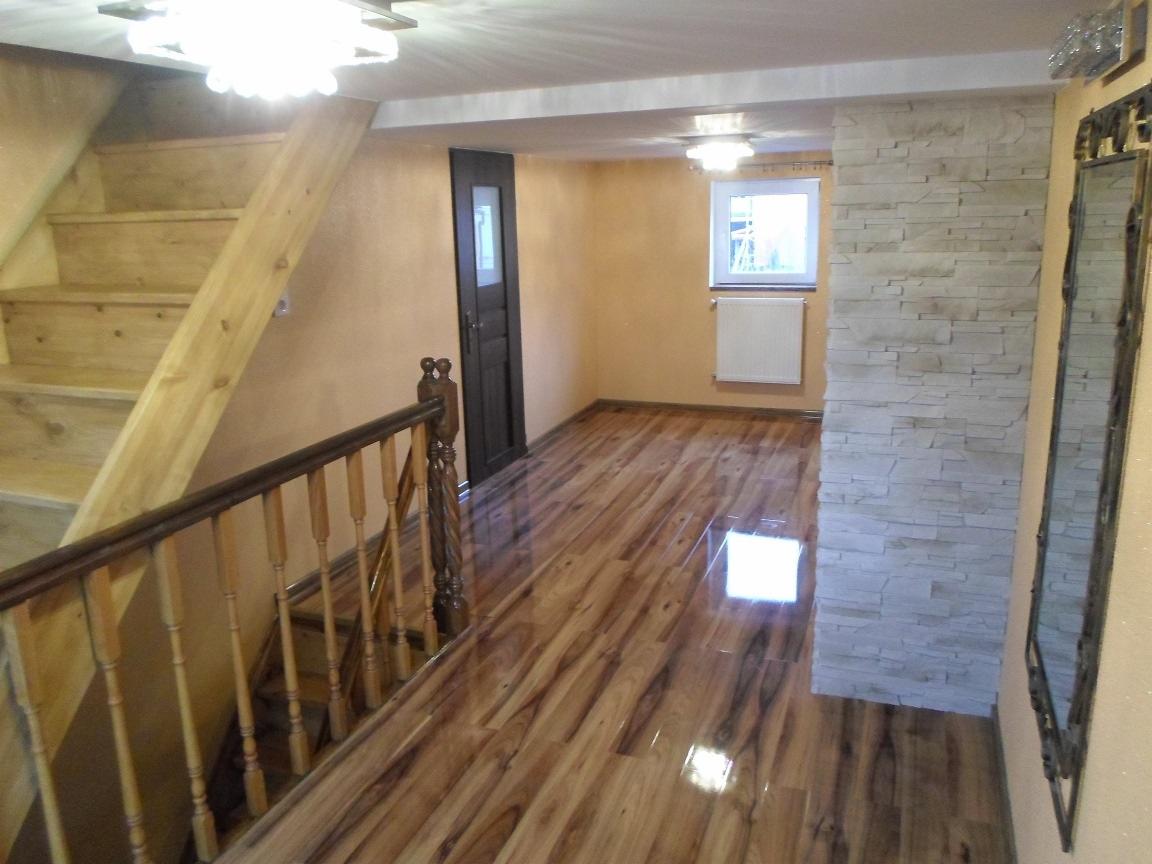 Wykończone wnętrze domu: drewniane schody z balustradą, lśniąca podłoga, ściana z kamienia dekoracyjnego, okno z grzejnikiem i ciemne drzwi. Ciepłe oświetlenie.