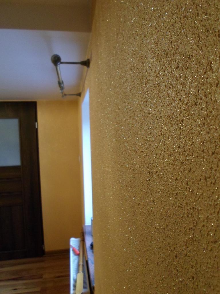 Błyszcząca, złota ściana z wyraźną fakturą w odcieniach beżu i brązu. Widoczne narzędzia malarskie przy ścianie,  oświetlenie i fragment drzwi.