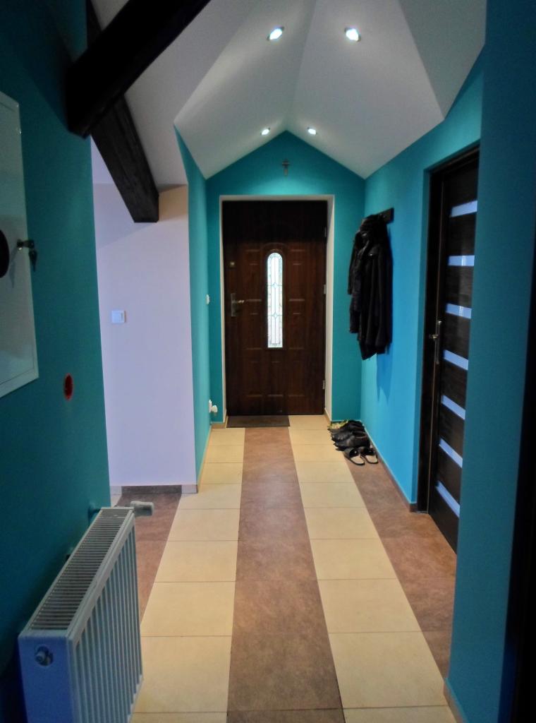 Turkusowy korytarz z beżowo-brązowymi płytkami podłogowymi, ciemnymi drzwiami wejściowymi z szybą i czarnymi drzwiami wewnętrznymi z poziomymi paskami.