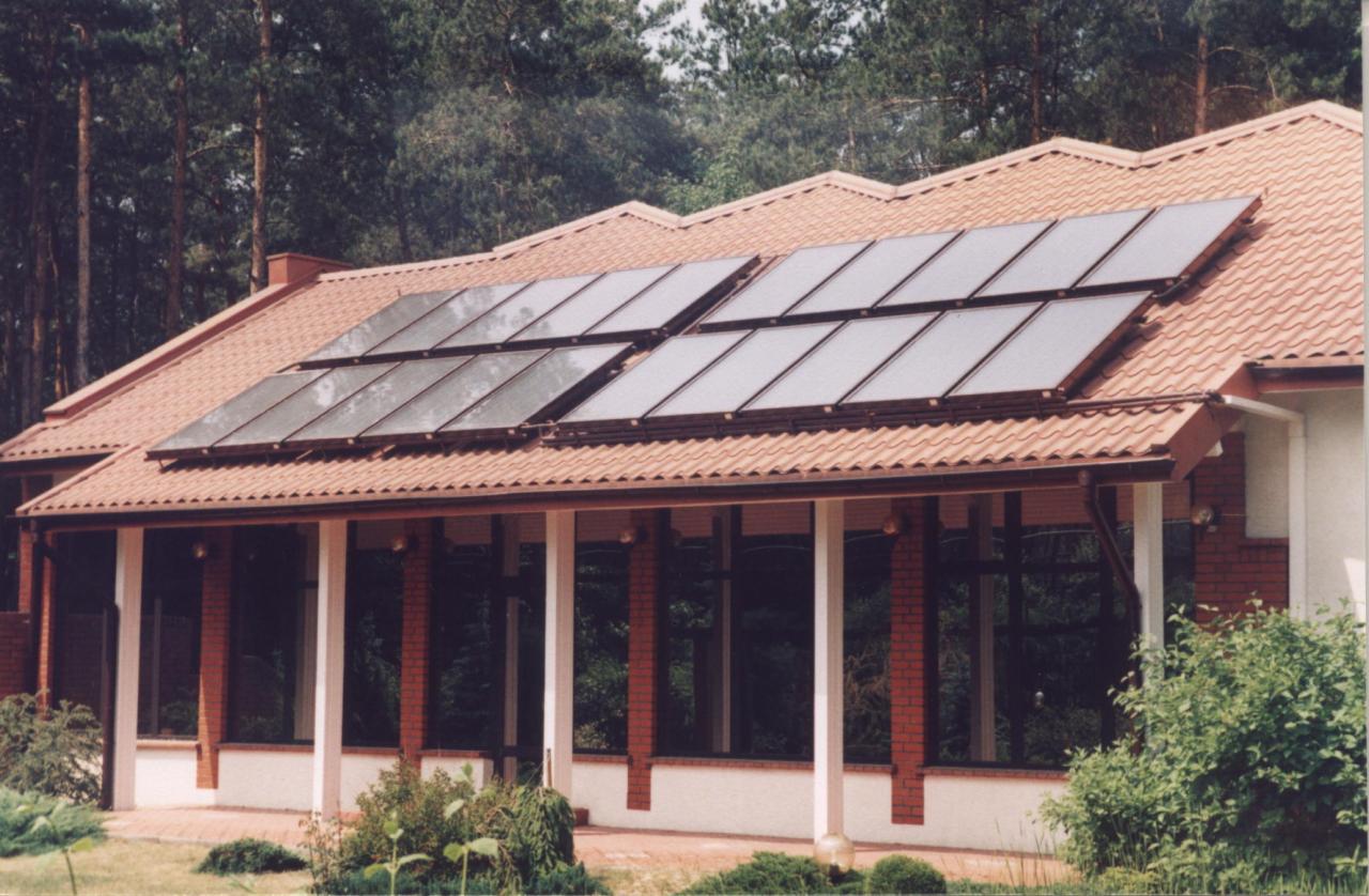 Dom jednorodzinny z brązową dachówką, na której zamontowane są panele fotowoltaiczne. Widok na część domu z podcieniem i kolumnami. W tle wysokie drzewa.