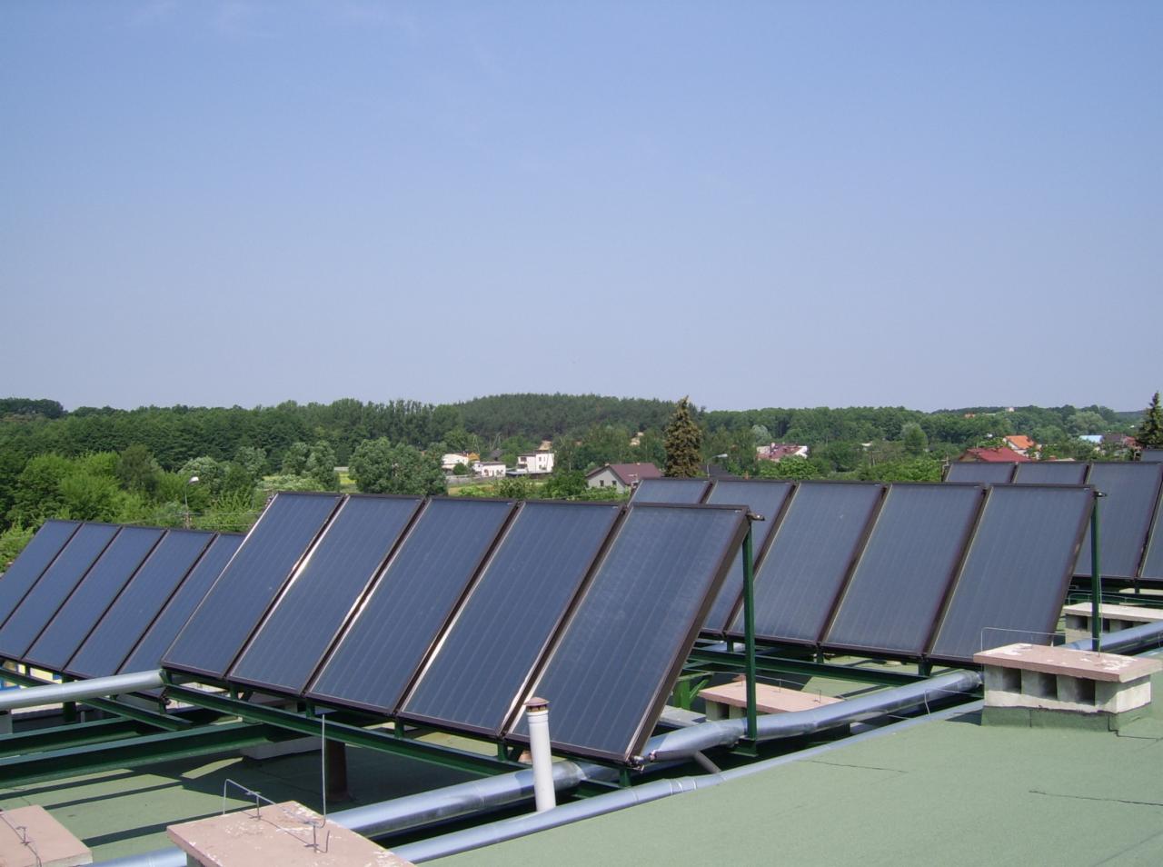 Rząd paneli słonecznych na zielonym dachu z widokiem na zalesiony teren. Metalowa konstrukcja wspiera panele, rury łączą system. Błękitne niebo w tle.