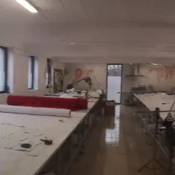 Wnętrze krojowni z długimi stołami, na których rozłożone są wykroje i materiały, a na ścianie wiszą szablony odzieżowe.