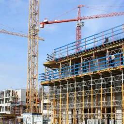 Wielopiętrowy budynek w trakcie budowy, widoczne rusztowania, szalunki i trzy żurawie wieżowe na tle błękitnego nieba.