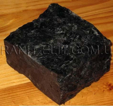 Czarny, surowy blok kostki granitowej o nieregularnych krawędziach, leżący na drewnianej powierzchni. Widoczne delikatne refleksy światła na powierzchni kamienia.
