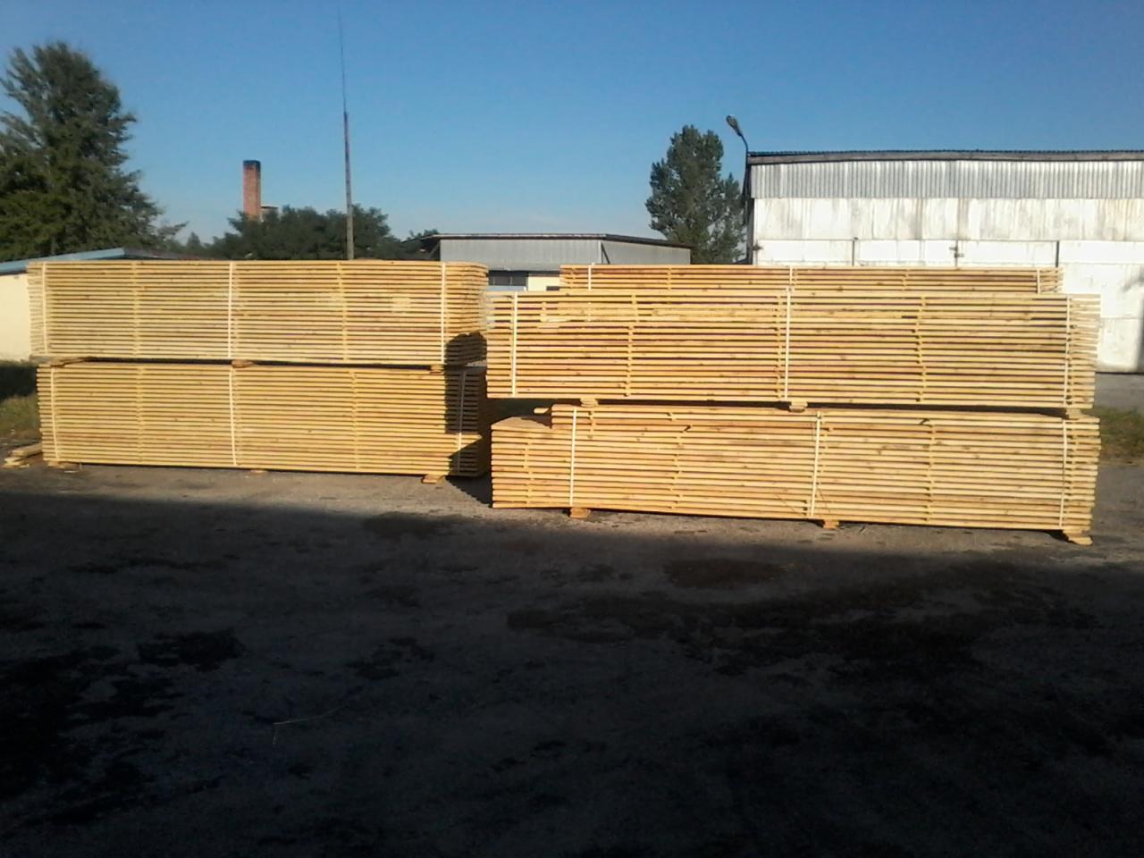 Stosy jasnej, świeżo obrzynanej deski sosnowej, zabezpieczone sznurkiem, ułożone na placu składowym z widocznym kominem w tle.