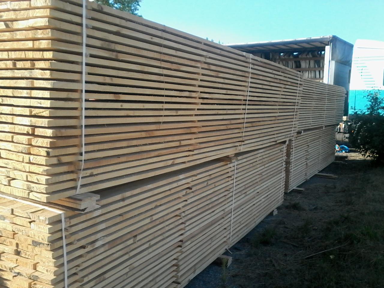 Stosy jasnej deski sosnowej obrzynanej, zabezpieczone białymi taśmami, ułożone na zewnątrz obok przyczepy ciężarówki w słoneczny dzień.