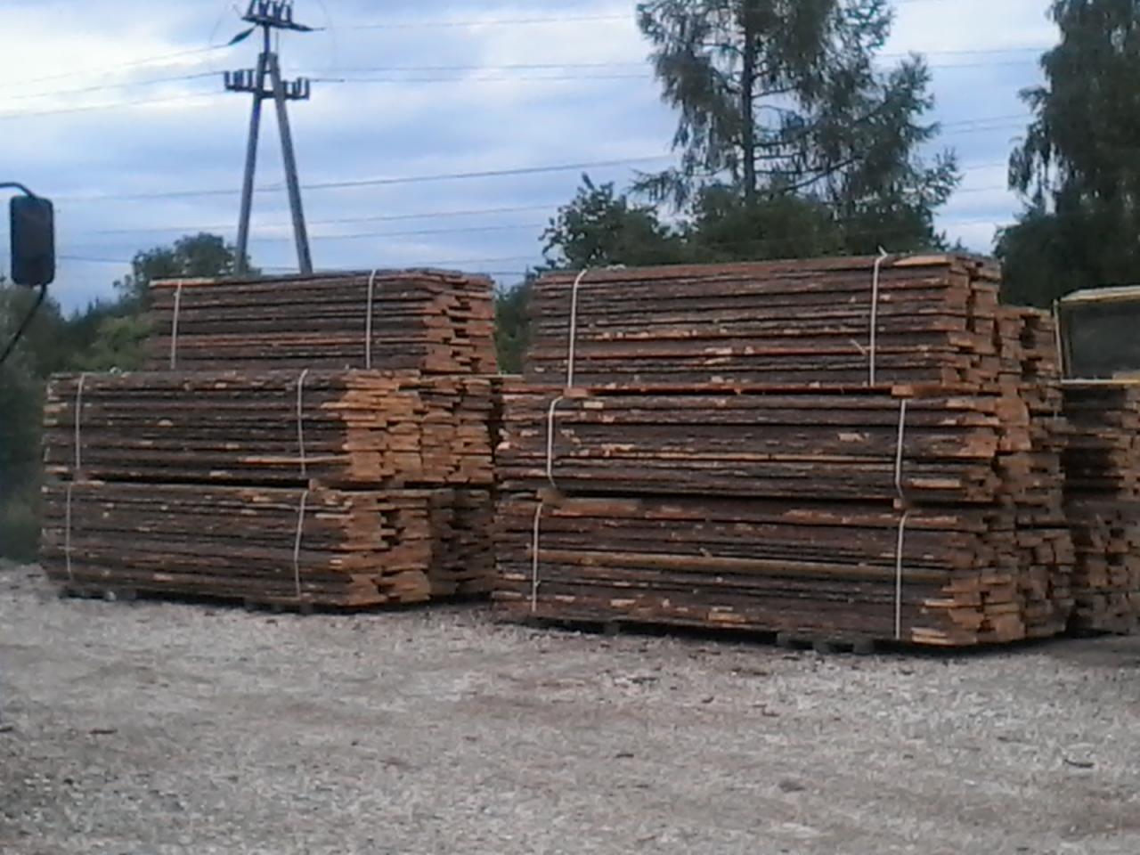 Stosy świeżo ściętej, suchej tarcicy sosnowej, związanej sznurkiem, ułożone na żwirowym placu składowym z widocznymi słupami elektrycznymi w tle.