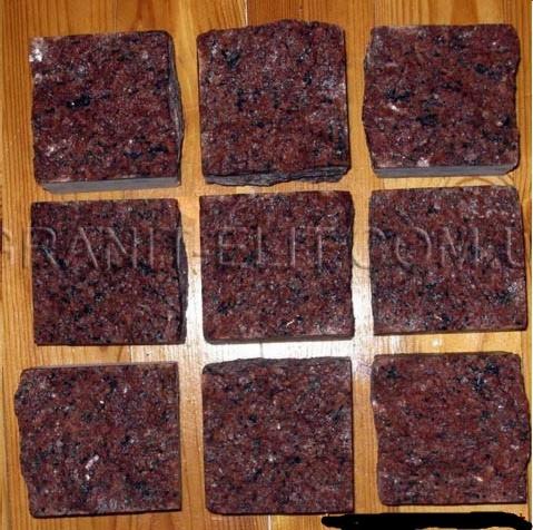 Dziewięć kostek granitowych w odcieniach czerwieni i brązu ułożonych na drewnianej powierzchni. Kostka granitowa oferowana w Woli Prażmowskiej.