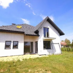 Niedawno wybudowany, biały dom jednorodzinny z szarą dachówką, widoczny z zewnątrz na tle błękitnego nieba i zielonego trawnika, z żółtą folią na dachu i opartymi o ścianę płytami izolacyjnymi.