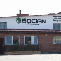 Budynek składu opału PHU Bocian z widocznym logo i adresem strony internetowej. Czerwona cegła, okna z kratami, szyld 'Autoryzowany Sprzedawca'.