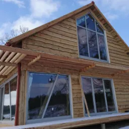 Surowy dom z bali w trakcie budowy, z widoczną drewnianą konstrukcją zadaszenia nad tarasem i dużymi oknami, na zimowym tle z lekkim śniegiem.