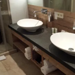 Nowoczesna łazienka z dwoma owalnymi umywalkami nablatowymi na ciemnym blacie, drewniana szafka z półkami na ręczniki, chromowana armatura i kabina prysznicowa w tle.