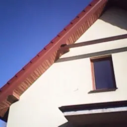 Czerwony dach z brązowymi obróbkami blacharskimi na jasnej elewacji domu, widok z dołu w słoneczny dzień. Widoczne okna i fragment balkonu.