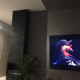 Częściowo wykończona ściana z dekoracyjnej, ciemnej mozaiki kamiennej, obok telewizor z włączonym obrazem. W rogu widoczna roślina doniczkowa.