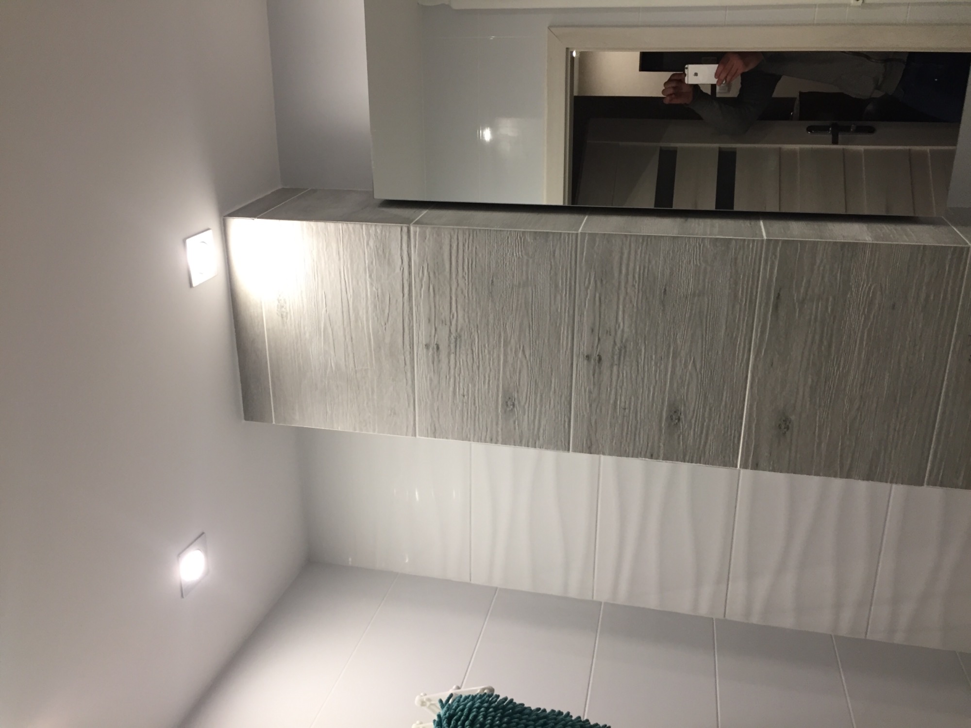 Wykończona ściana w łazience z szarymi płytkami imitującymi drewno u góry i białymi płytkami poniżej, odbicie fotografa z telefonem w lustrze.