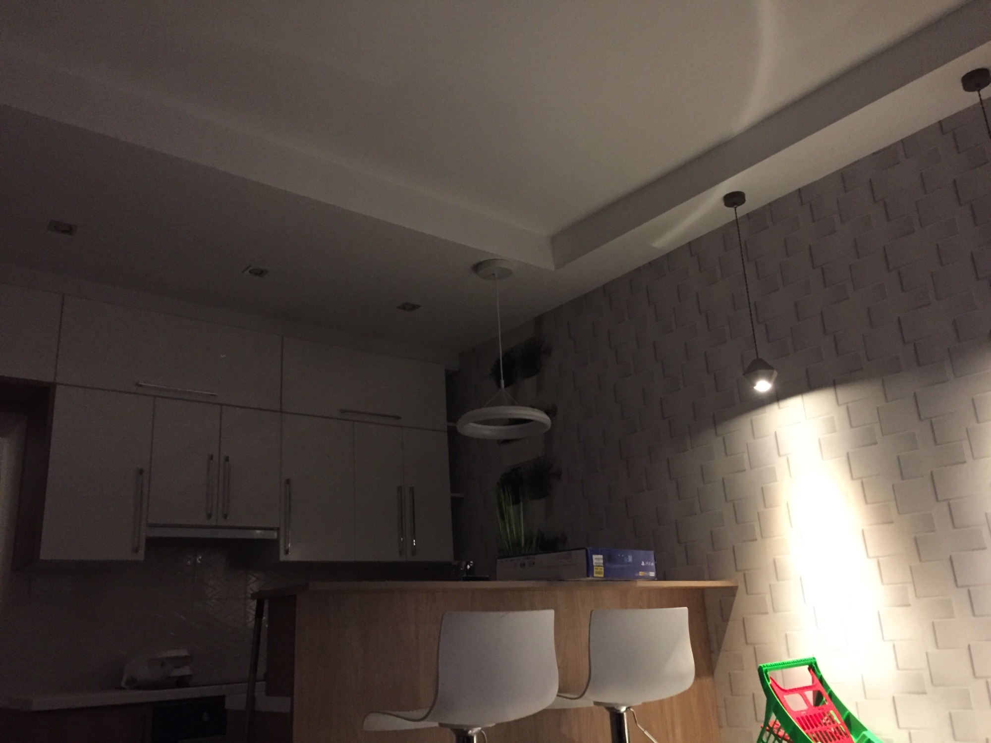 Nowoczesna kuchnia z białymi szafkami, drewnianym blatem, dwoma białymi hokerami i oświetleniem punktowym oraz dekoracyjną ścianą z efektem 3D, częściowo widoczny wózek na zakupy dla dziecka.