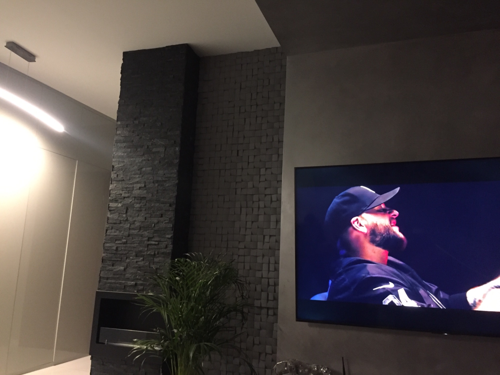 Częściowo wykończona ściana z dekoracyjnej, ciemnej mozaiki kamiennej, obok telewizor z włączonym obrazem. W rogu widoczna roślina doniczkowa.