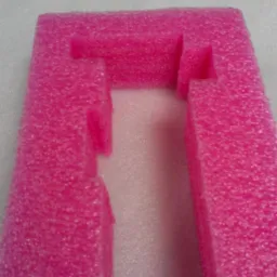 Różowa pianka polietylenowa z wyciętymi kształtami, tworząca ramkę z wgłębieniami. Detal opakowania ochronnego na jasnym tle, widok z góry.