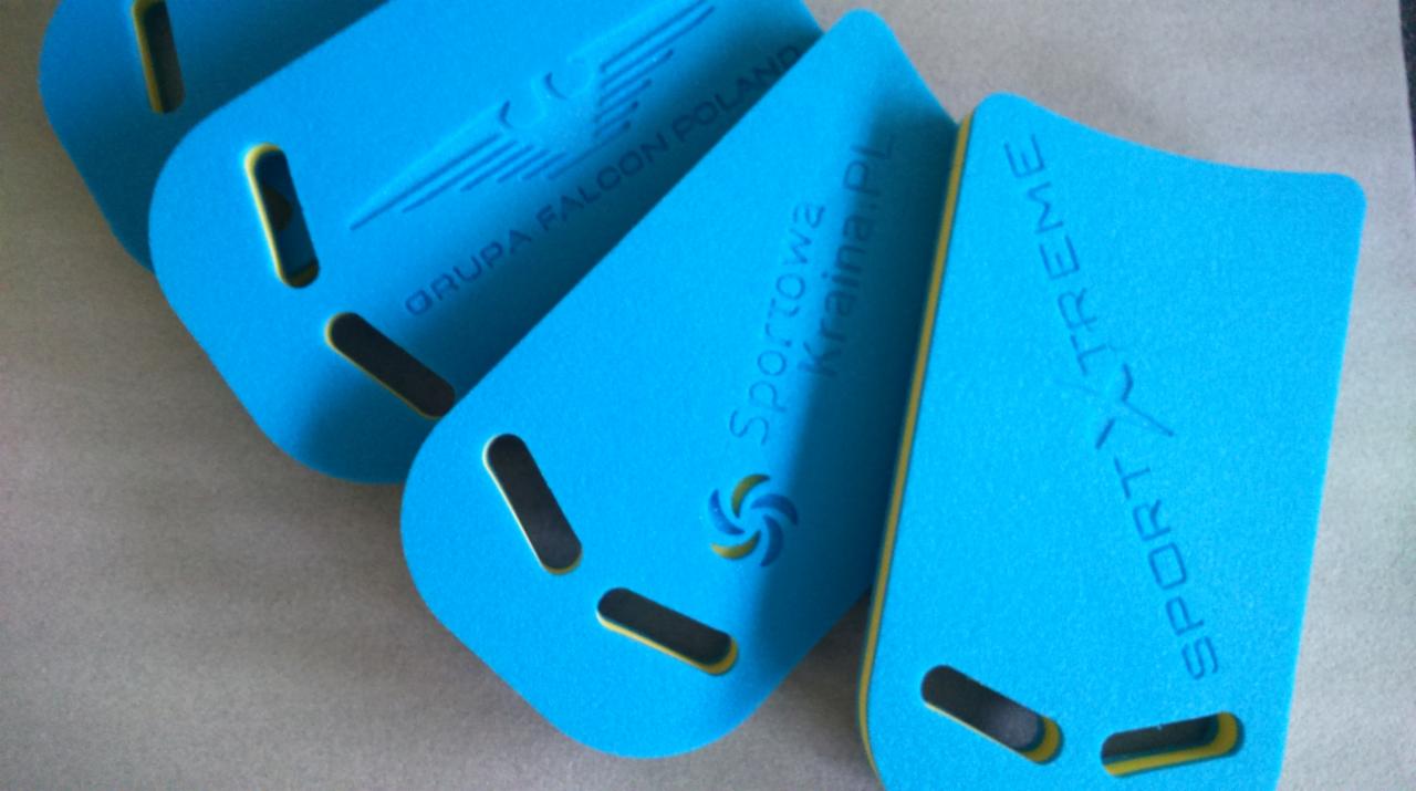 Błękitne deski do pływania z żółtymi krawędziami i wyciętymi otworami, ułożone pod kątem, z nadrukami firmowymi. Detaliczne ujęcie, ukazujące strukturę materiału.