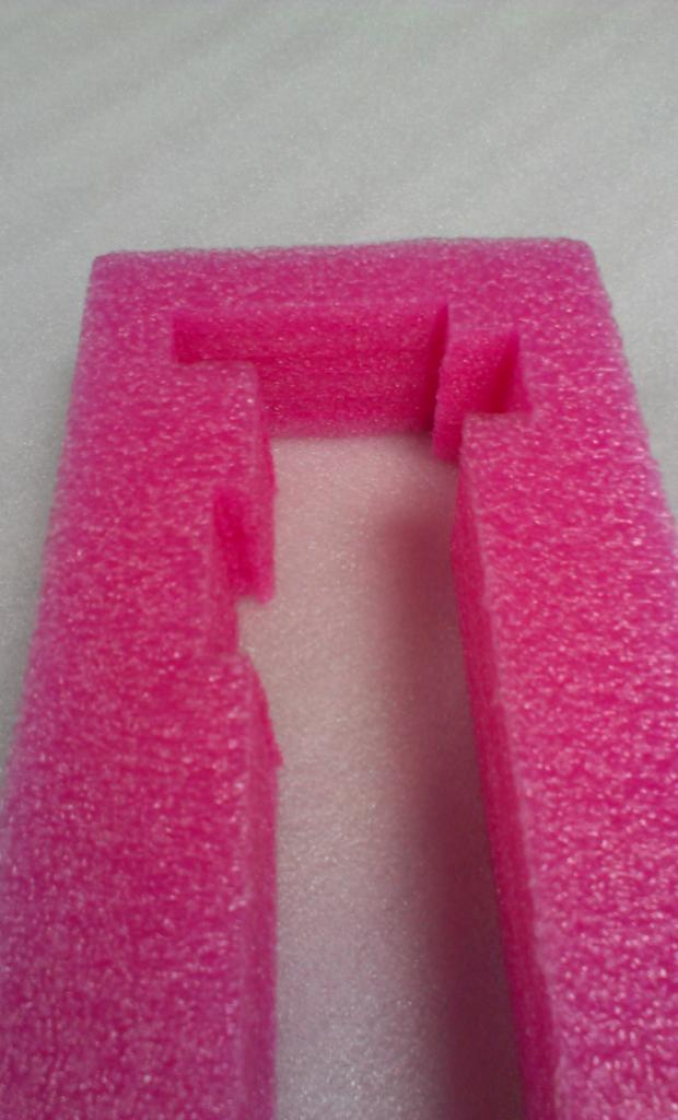 Różowa pianka polietylenowa z wyciętymi kształtami, tworząca ramkę z wgłębieniami. Detal opakowania ochronnego na jasnym tle, widok z góry.