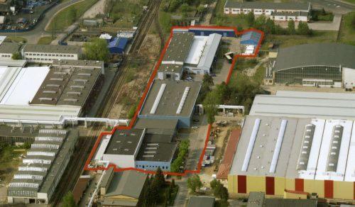 Fabryka opakowań z lotu ptaka, obrysowana czerwoną linią, z widocznymi halami produkcyjnymi i torami kolejowymi w pobliżu. Ujęcie z góry.