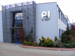 Budynek firmy PJ z dużym logo na elewacji. Szary budynek z nowoczesnym oknem i zielenią wokół. Widok z zewnątrz, w dzień.