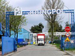 Brama wjazdowa do fabryki z logo 'P' i napisem 'PRZEDSIĘBIORSTWO'. Czerwono-biała ciężarówka wjeżdża na teren zakładu. Widoczne znaki drogowe i budynki.