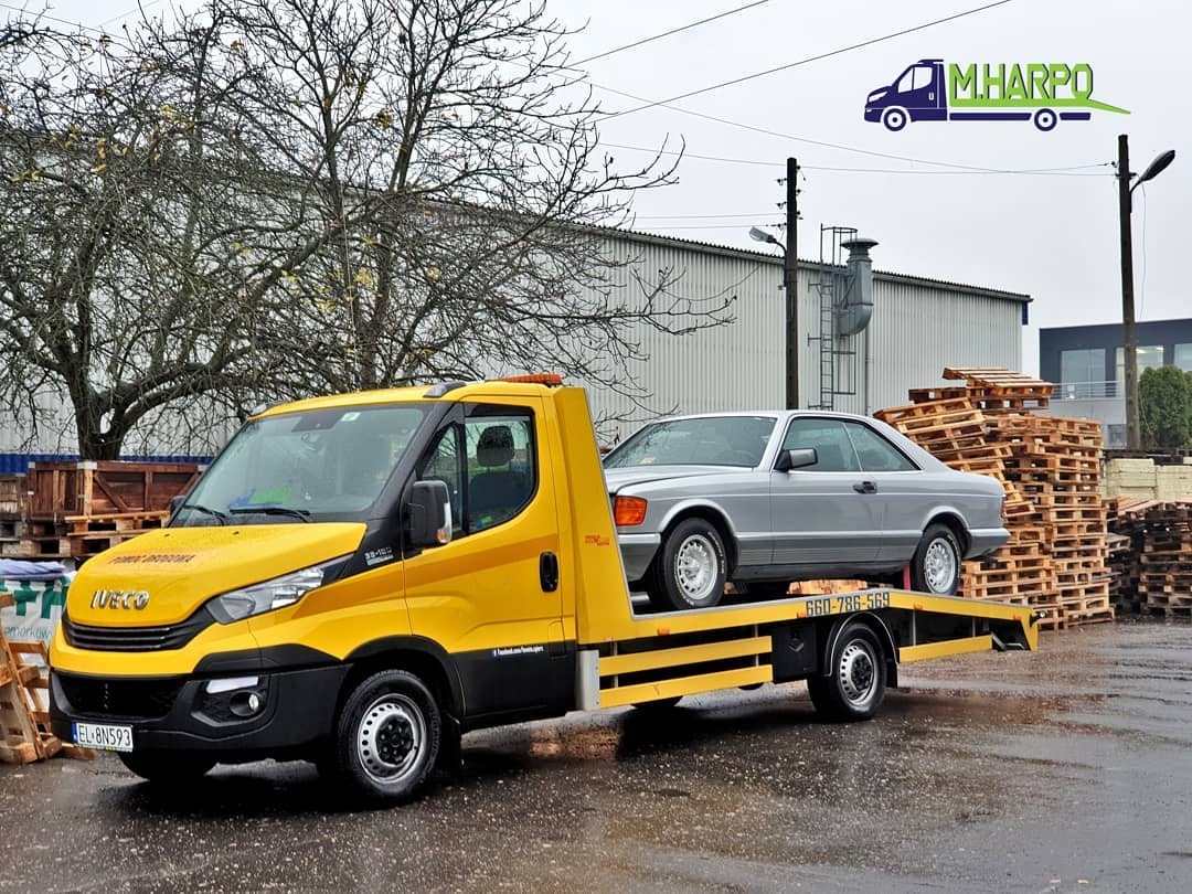 Żółta laweta Iveco z szarym Mercedesem na platformie, na tle magazynu i stosu drewnianych palet.