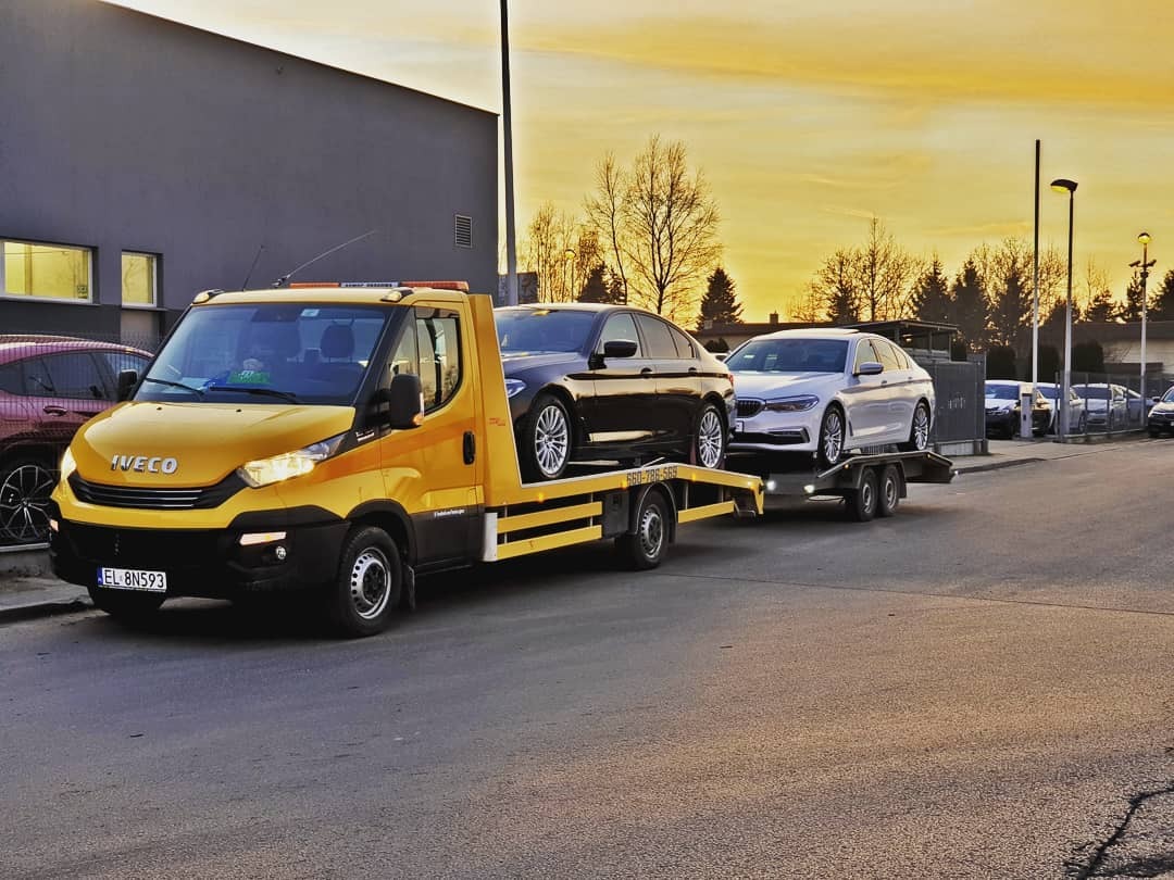 Żółta laweta Iveco z czarnym i białym BMW na platformie, zaparkowana na ulicy przy szarym budynku, w tle zachodzące słońce.