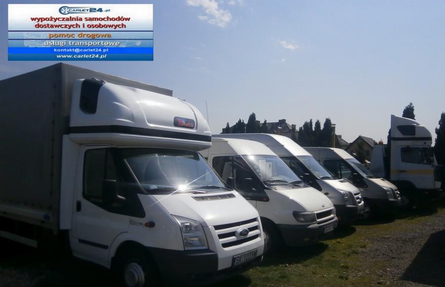 Rząd białych busów dostawczych marki Ford Transit, ustawionych na trawiastym placu, z logo firmy Carlet24 w lewym górnym rogu i budynkami w tle.