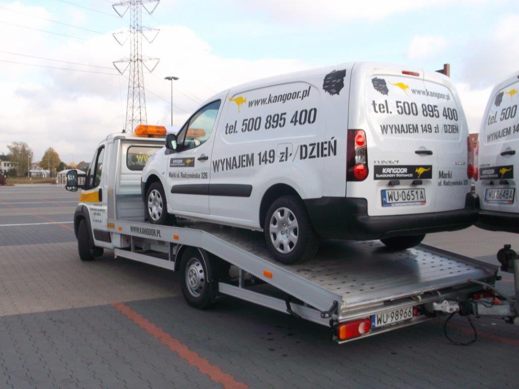 Laweta z pomarańczowym światłem ostrzegawczym przewozi biały samochód dostawczy z napisem 'Wynajem 149 zł / dzień' na parkingu z szarą kostką brukową, w tle słup wysokiego napięcia.